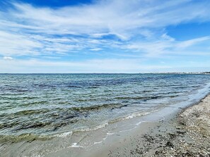 A beach with clear water, pebbly shore, and a distant cityscape.