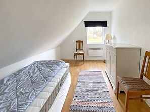 Quarto pequeno e bem iluminado, com cama, cadeira, escrivaninha e janela com cortina preta.