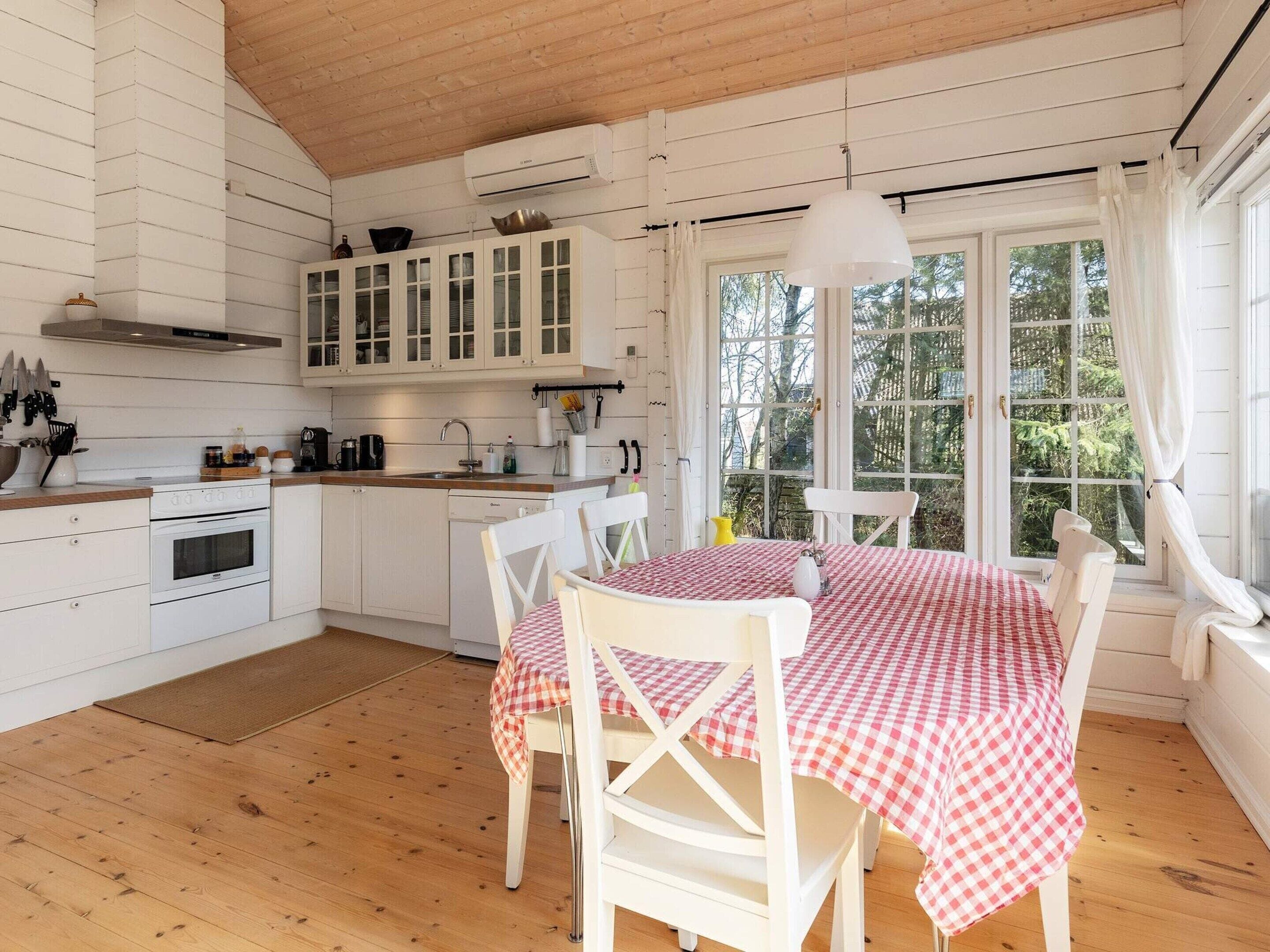 Une cuisine avec des meubles blancs, une cuisinière et un évier. On y trouve une table à manger recouverte d’une nappe à carreaux et entourée de chaises blanches.