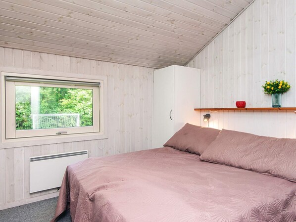 A bedroom with a large window, a bed with pink bedding, a white wardrobe, and a small shelf with a vase of flowers.