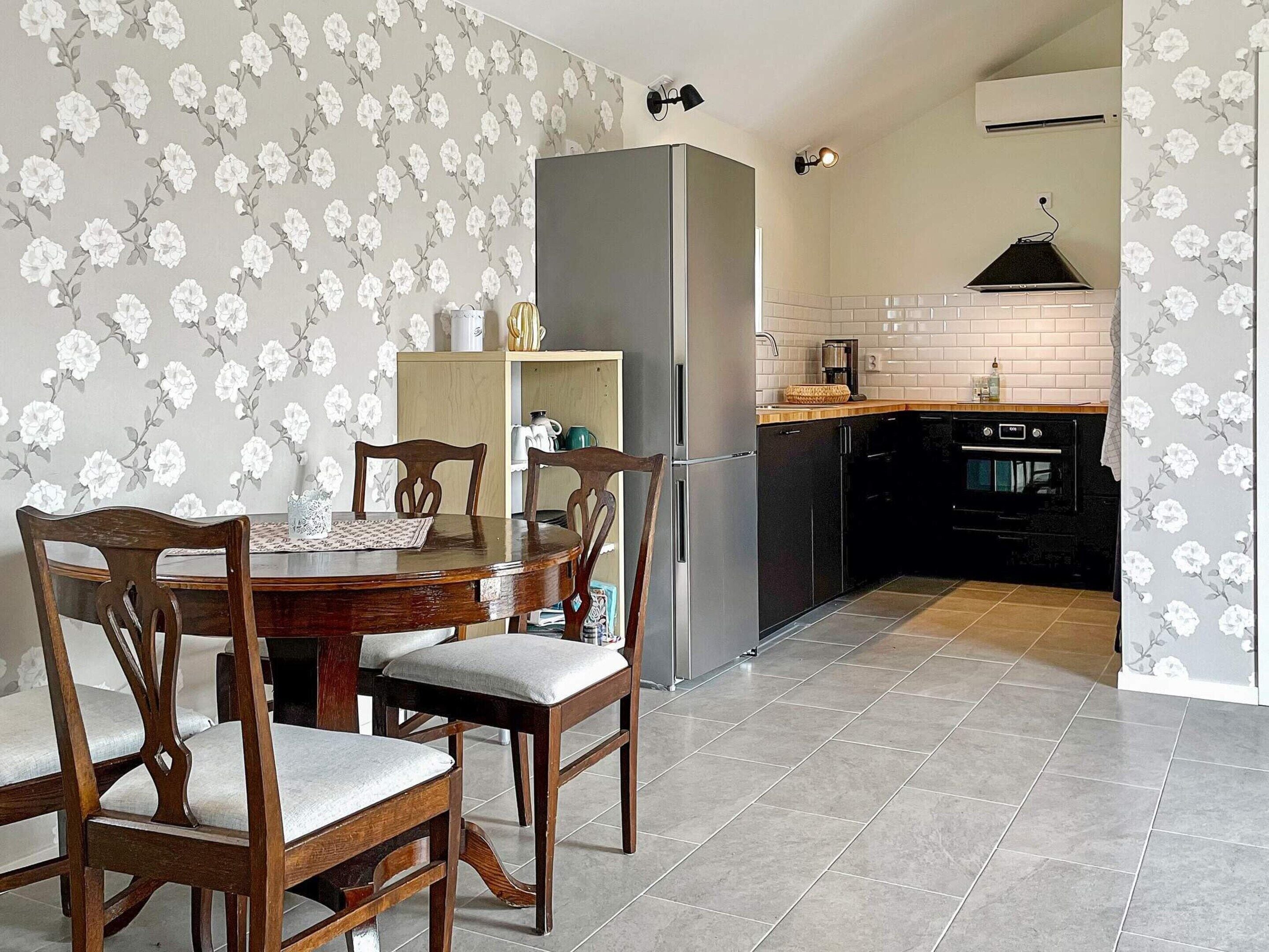 A dining area with a wooden table and chairs, a refrigerator, and a kitchen with a tiled backsplash and black appliances.