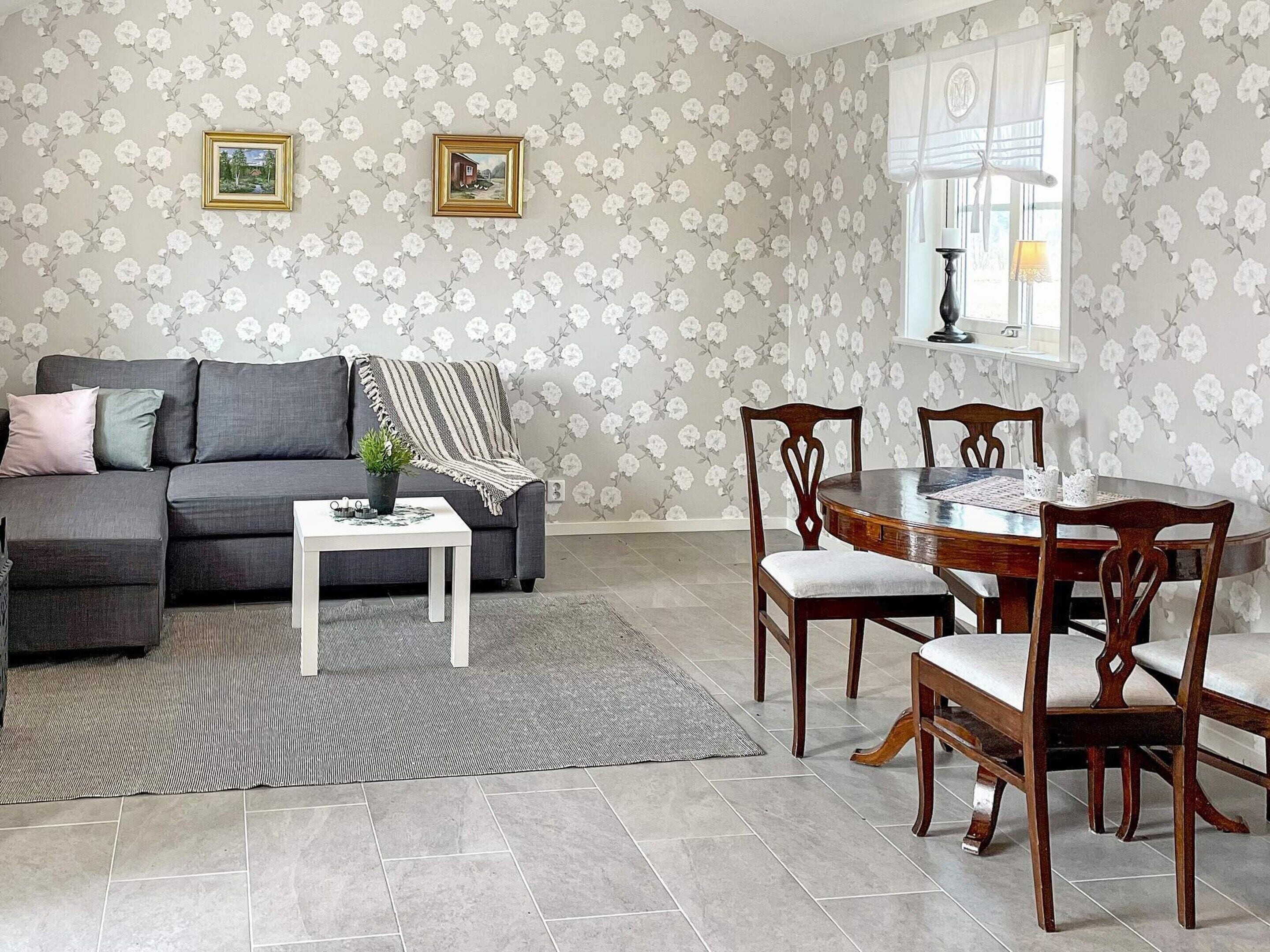 A living room with a grey sofa, a small white table, and a wooden dining table with chairs. There are floral wallpaper and two framed pictures on the wall.