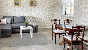 A living room with a grey sofa, a small white table, and a wooden dining table with chairs. There are floral wallpaper and two framed pictures on the wall.