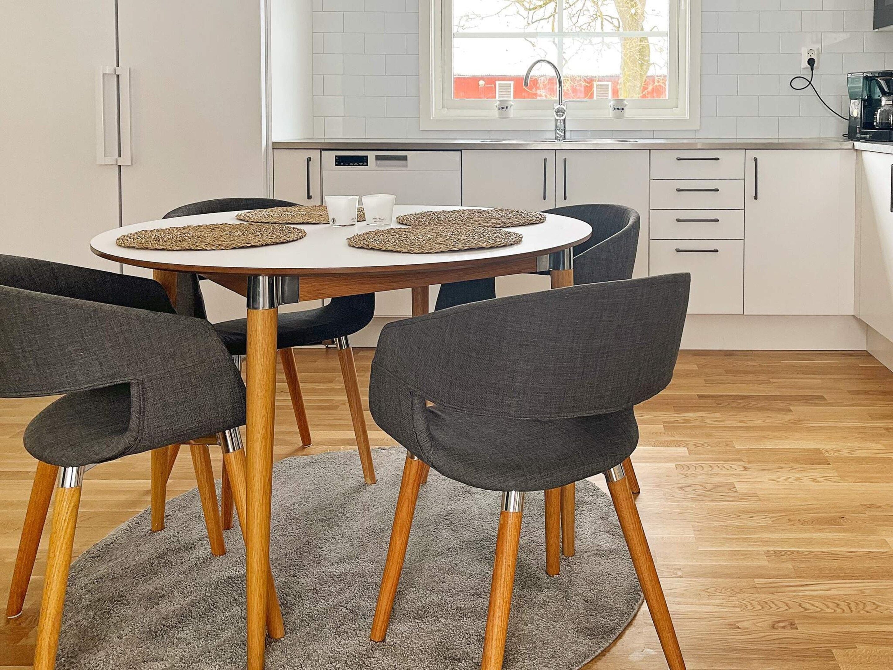 A modern kitchen with white cabinets, a round dining table with four chairs, and a window with a view of trees.
