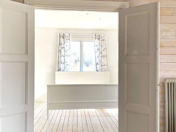 Um quarto com radiador branco, janela com cortinas estampadas e assoalho de madeira.