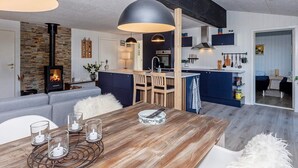 A modern kitchen with a central island, a dining area with a wooden table and chairs, and a cozy living space with a sofa and a fireplace. - 6 Person Holiday Home in Farso (Farso)