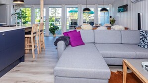 A modern living room with a grey sectional sofa, wooden dining table, and a kitchen area with a built-in island. - 6 Person Holiday Home in Farso (Farso)