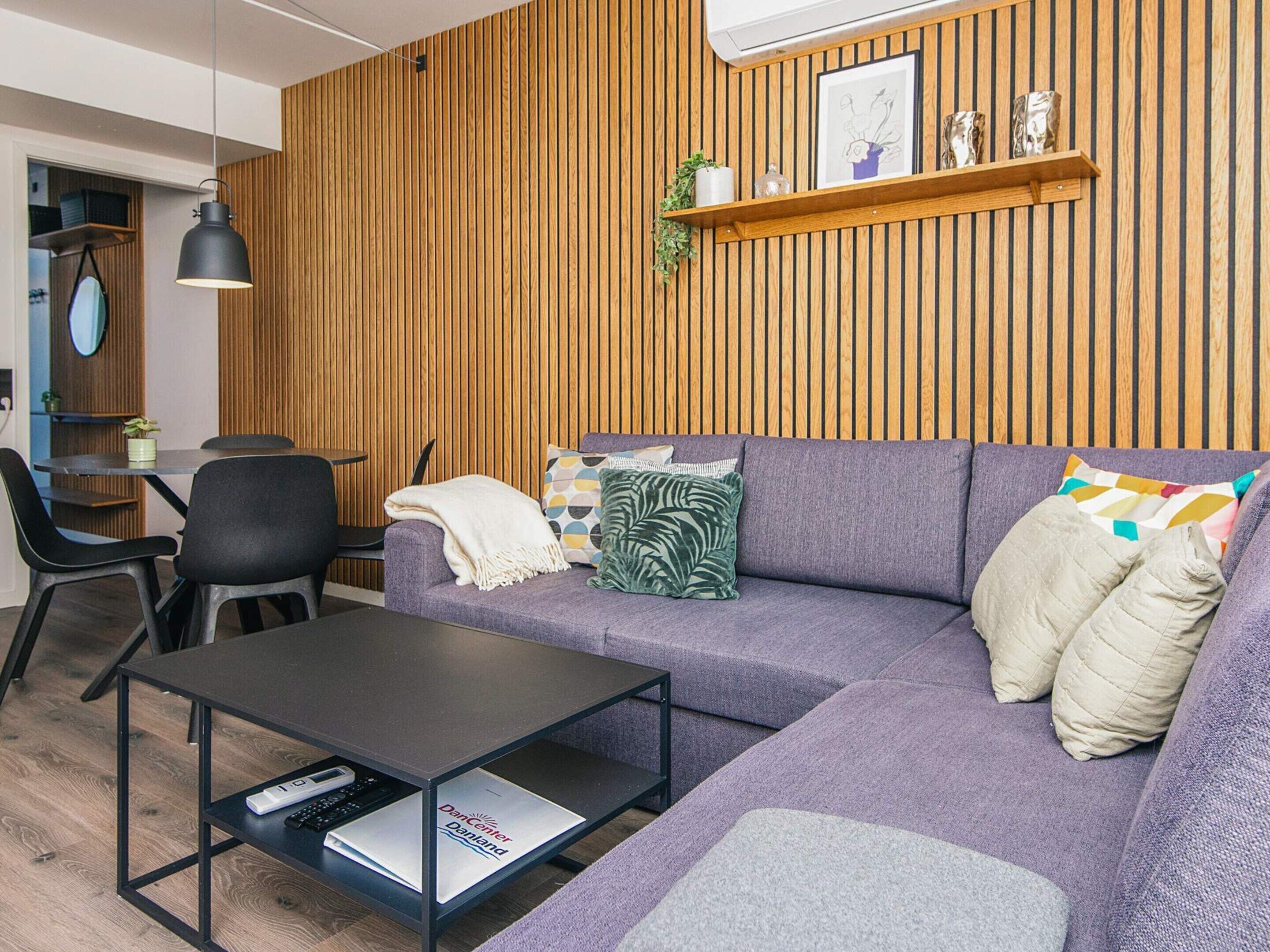 A modern living room with a wooden panel wall, a purple sofa, a black coffee table, and a small dining area with a table and chairs.