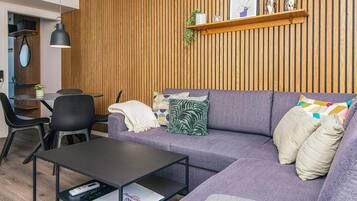 A modern living room with a wooden panel wall, a purple sofa, a black coffee table, and a small dining area with a table and chairs.