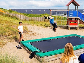 A trampoline on a sandy surface with a wooden fence and a blue playhouse in the background. - 4 Star Holiday Home in Ringkobing (Ringkobing)