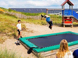 A trampoline on a sandy surface with a wooden fence and a blue playhouse in the background.