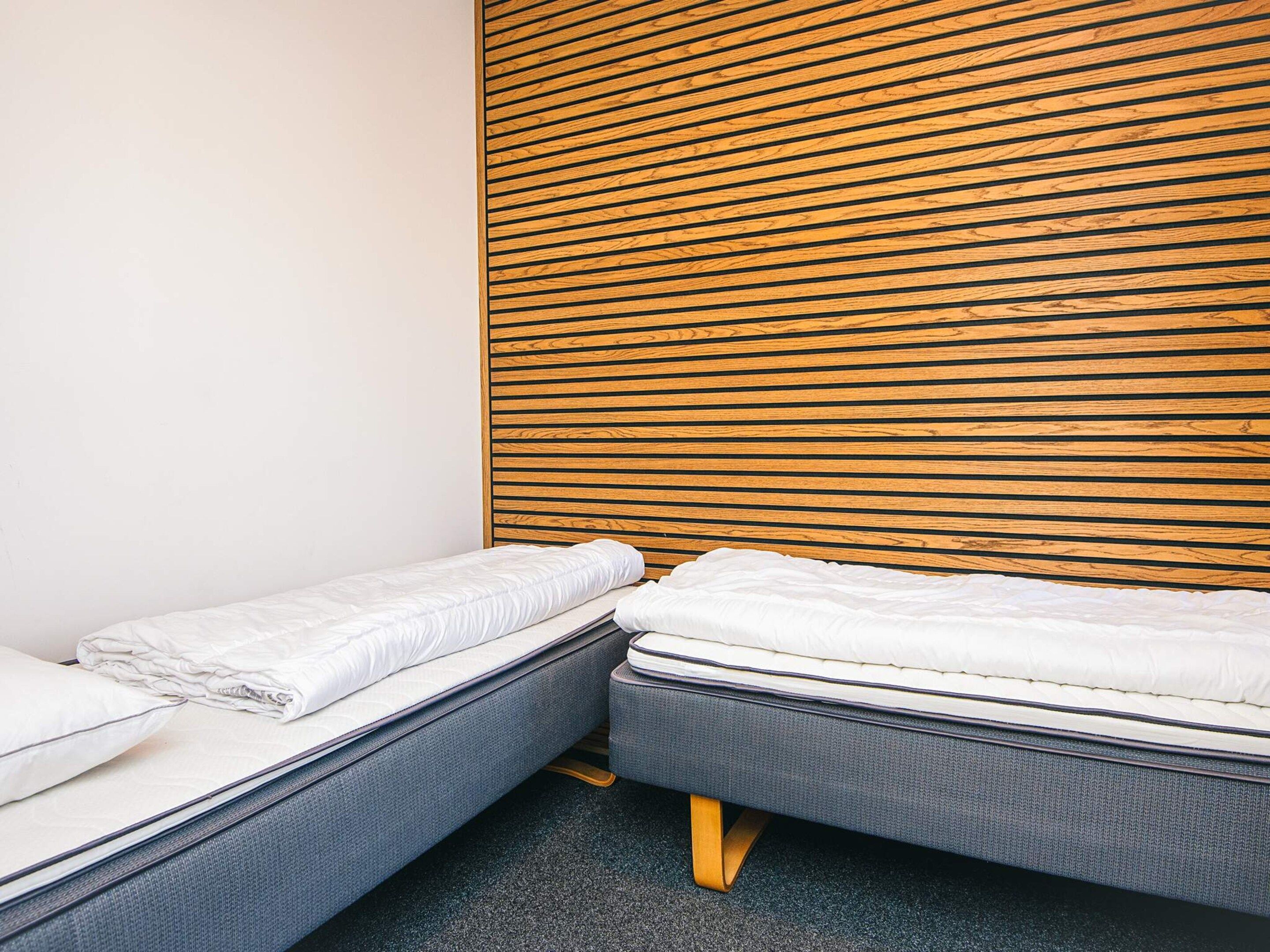 A room with two single beds, a wooden slatted headboard, and a plain white wall.