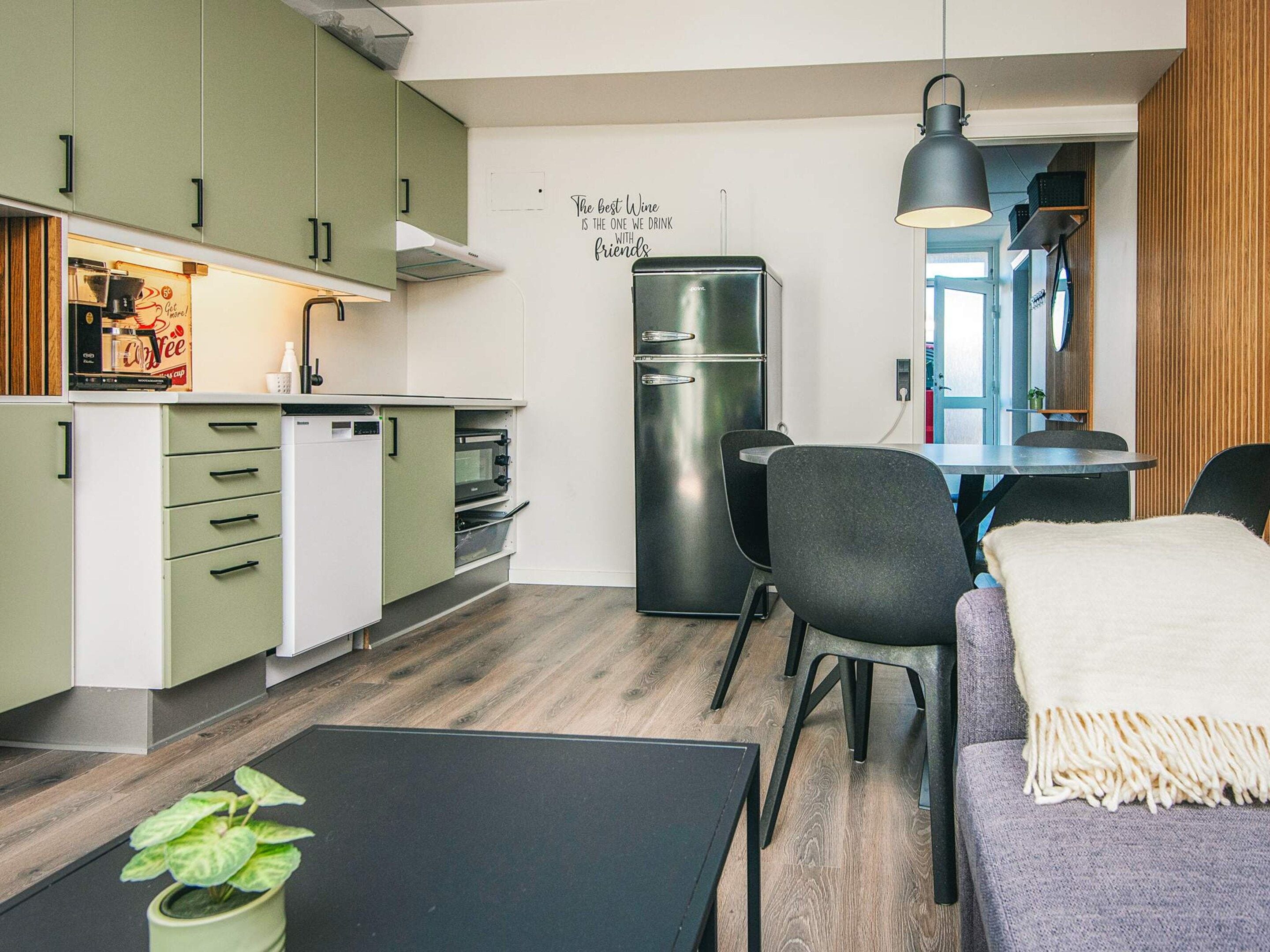 A compact kitchen with a refrigerator, microwave, and coffee machine, a dining area with a table and chairs, and a wall with a motivational quote.