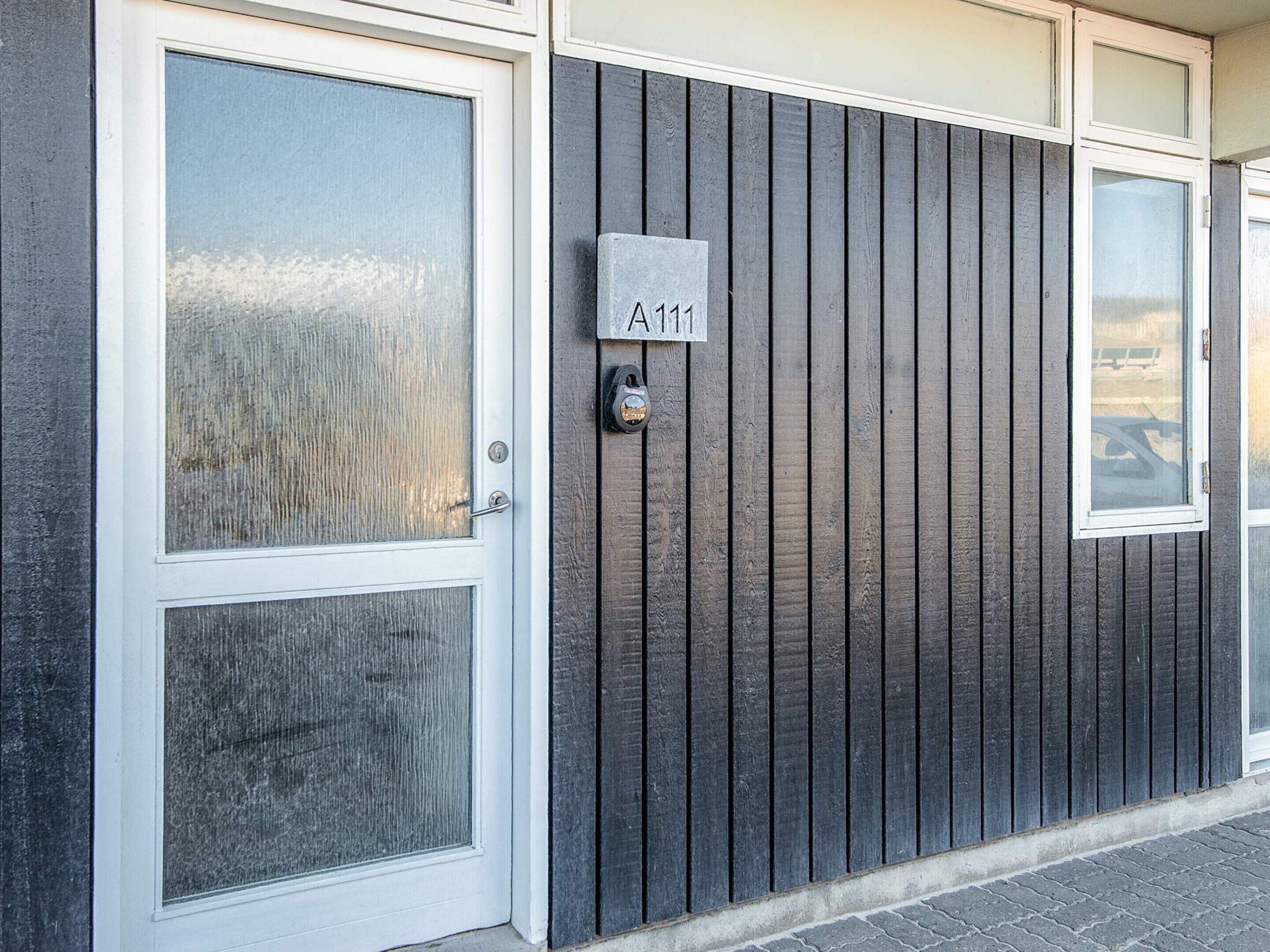 Fachada de edificio con paneles exteriores de madera oscura, una puerta blanca con un panel de vidrio y un buzón con placa de número de casa.