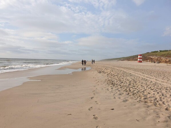 A wide sandy beach with footprints leading to the water, a red and white lifeguard tower, and a grassy hill in the background. - 4 Star Holiday Home in Ringkobing (Ringkobing)