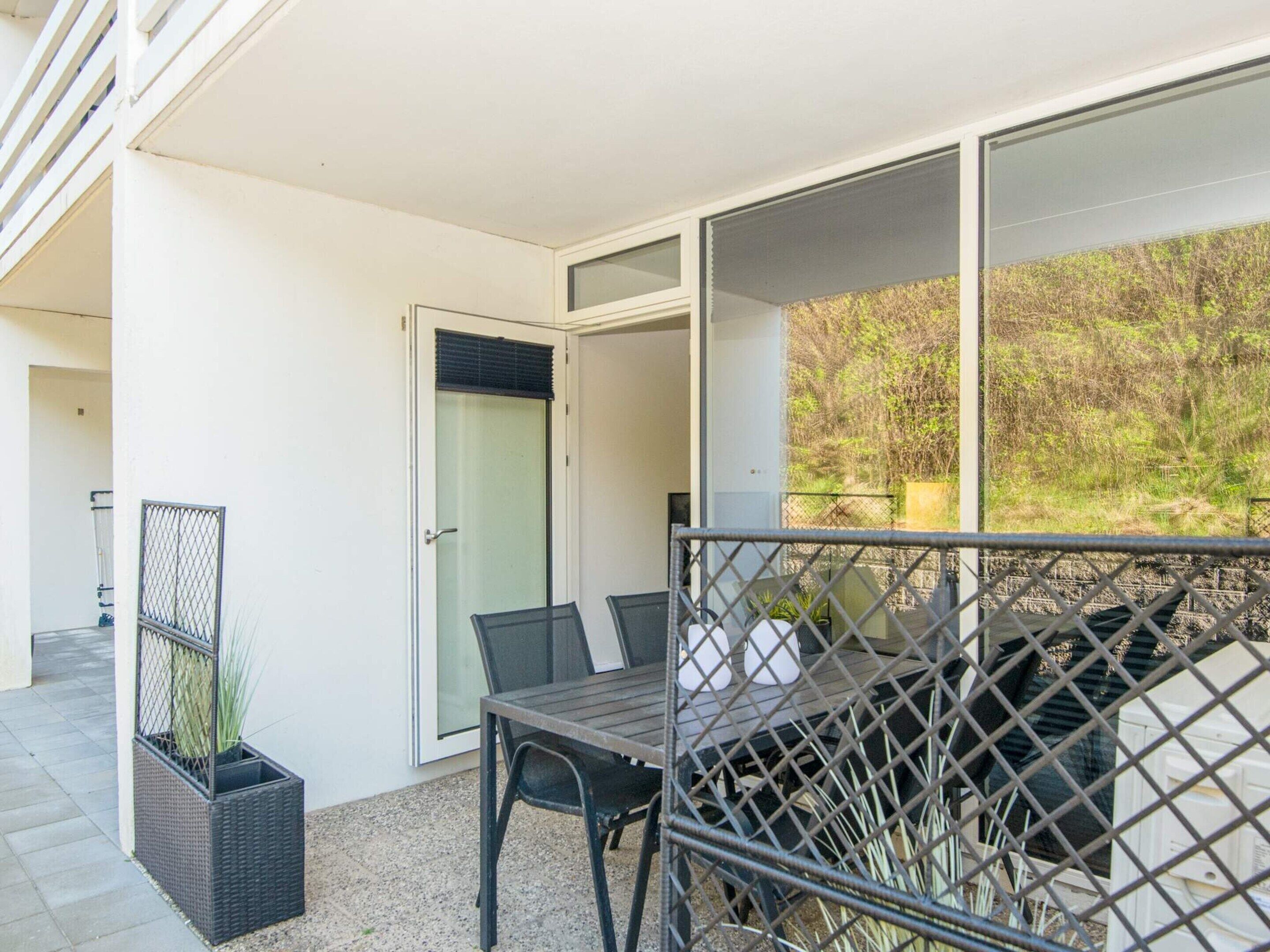 Un balcón con mesa y sillas, una puerta con panel de vidrio y vista a la vegetación.