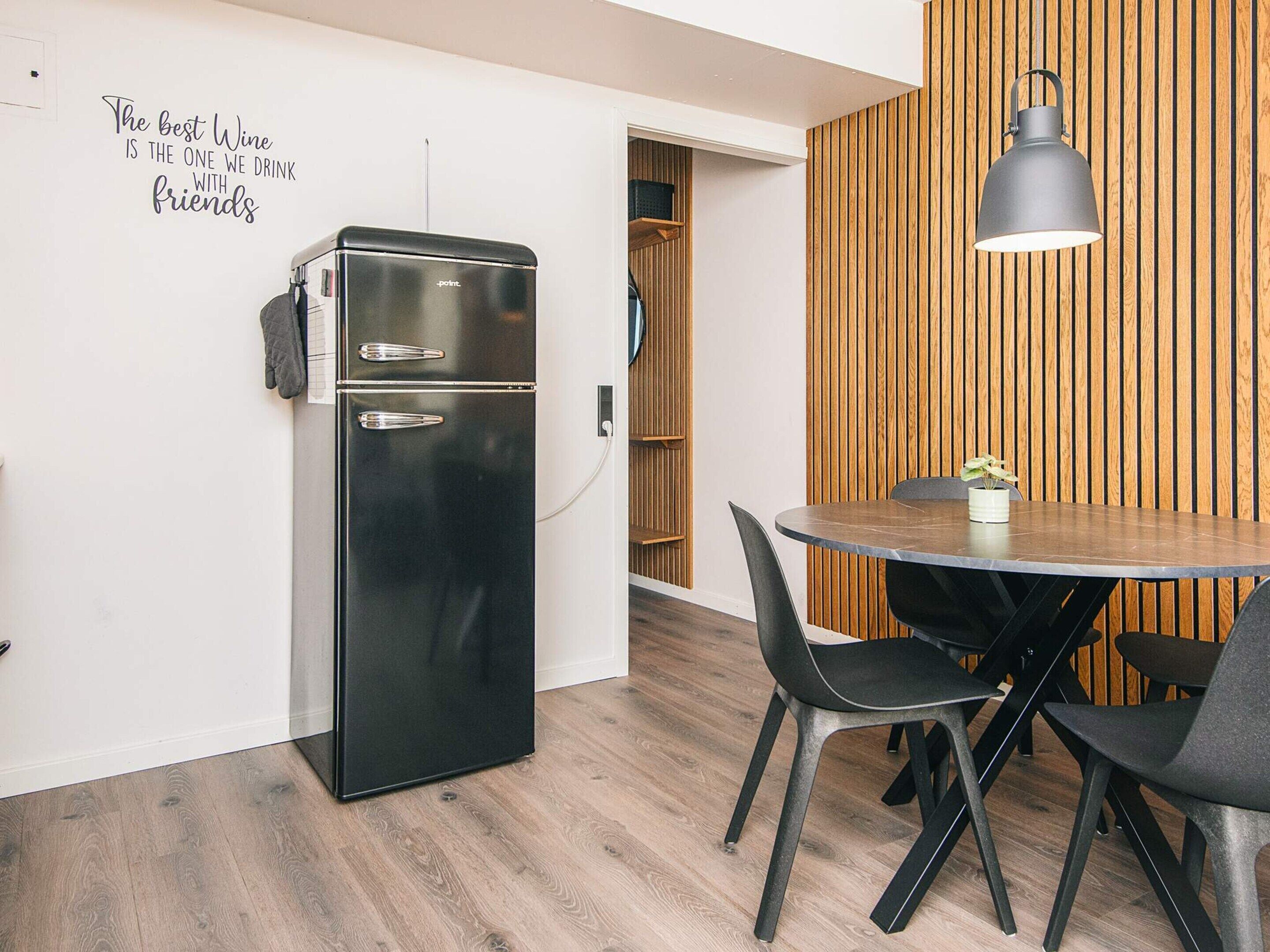 A compact kitchen with a black refrigerator, a wooden wall panel, a dining table with chairs, and a pendant light.