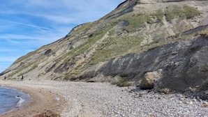 Un paisaje costero con una playa pedregosa, un acantilado con formaciones rocosas estratificadas y un cielo despejado.