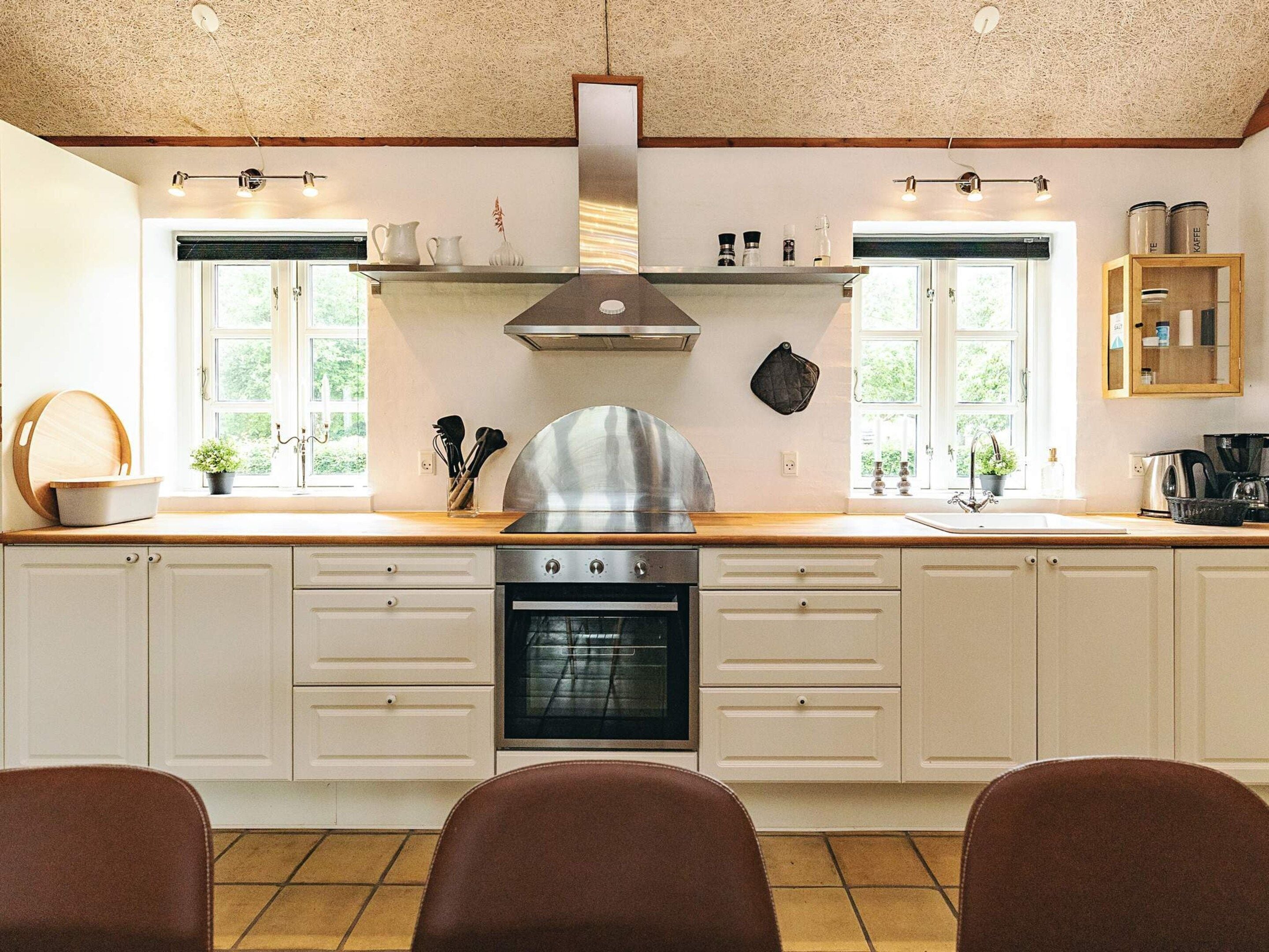 A kitchen with a stove, oven, and a range hood. There are two windows with curtains, a sink, and a countertop with various items.