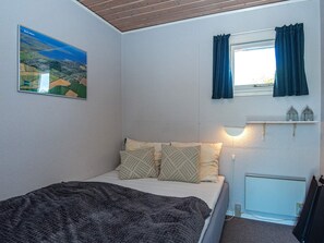 Un dormitorio con cama, ventana con cortinas, estante con dos faroles y un cuadro en la pared.