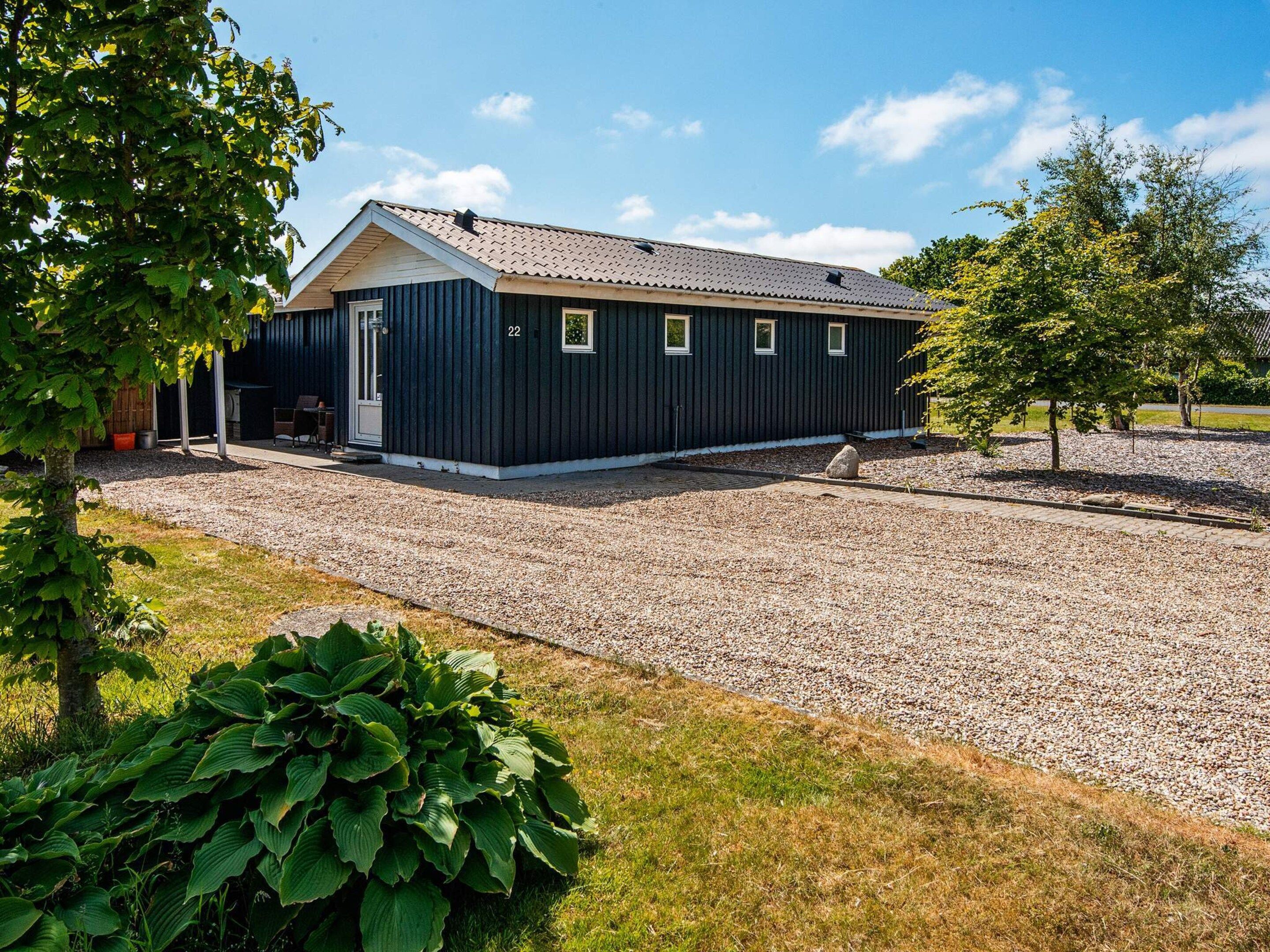 En etagers bygning med mørk facade, grusvej og en lille have med et træ og en stor, grøn plante.