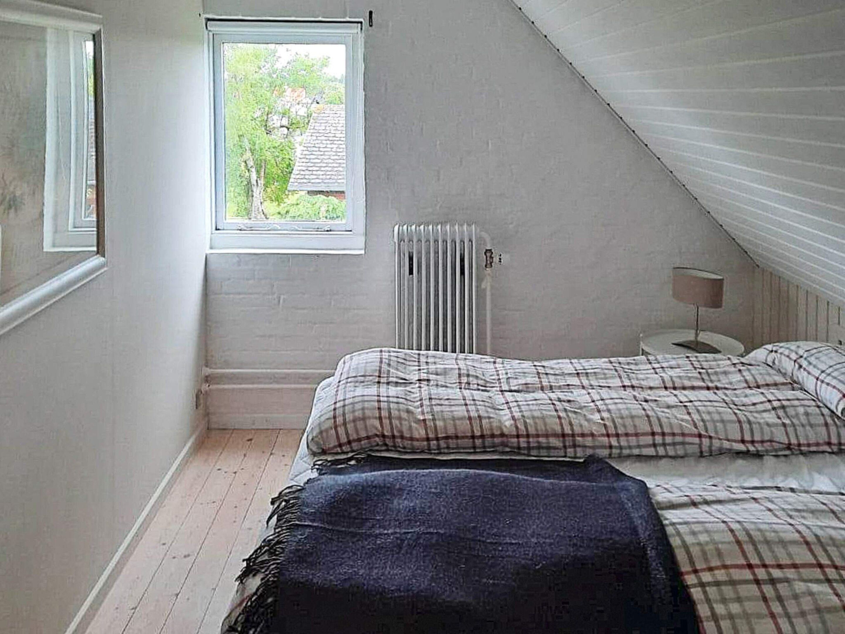 A bedroom with a sloped ceiling, a window, a radiator, a bedside table with a lamp, and a bed with plaid bedding.