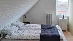 A bedroom with a sloped ceiling, a bed with patterned bedding, a bedside table, a radiator, and a window with a view of buildings. - 6 Person Holiday Home in Bindslev (Bindslev)