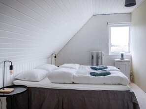 Une chambre avec un plafond en pente, un radiateur, une fenêtre et un lit avec des oreillers et une couverture.
