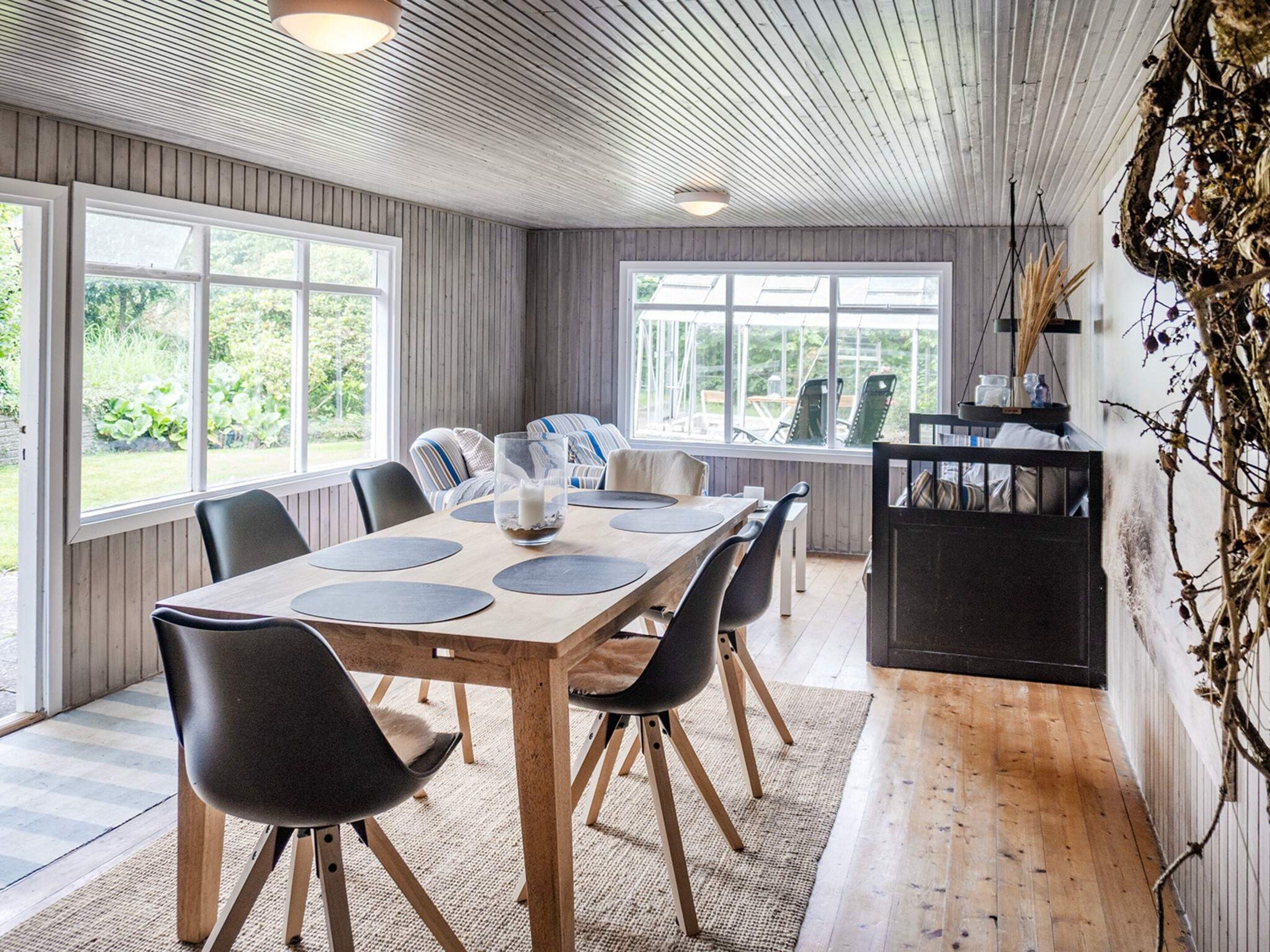 Un espace repas avec une table en bois, des chaises noires et une vue sur l’extérieur grâce à de grandes fenêtres.