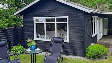 Una pequeña estructura de madera oscura con una ventana y una puerta, rodeada de césped y una silla de metal negro.