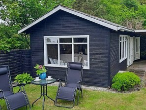 A small, dark-colored wooden structure with a window and a door, surrounded by a grassy area and a black metal chair. - 6 Person Holiday Home in Bindslev (Bindslev)