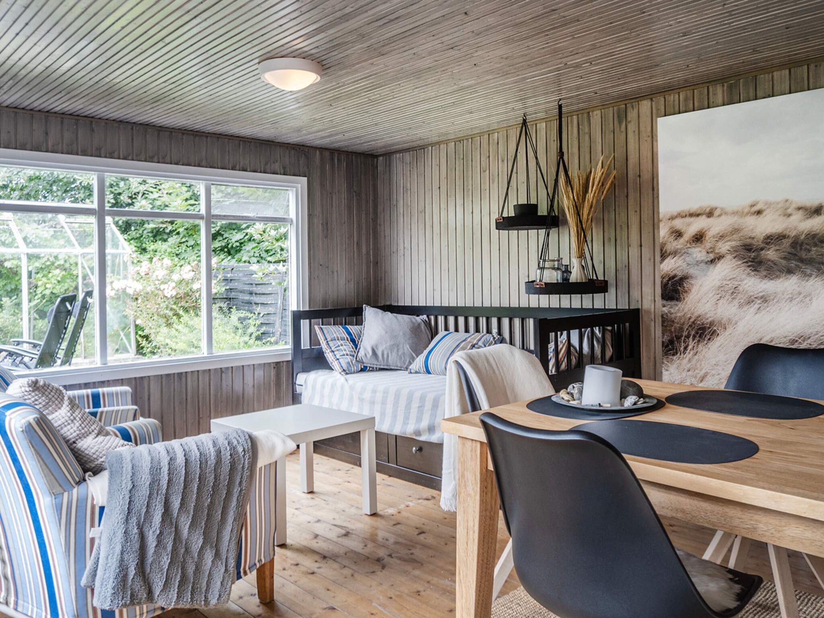 A cozy room with a wooden ceiling, a large window, a bed with striped bedding, a dining table with chairs, and a painting on the wall.