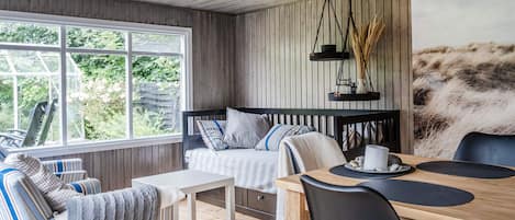 A cozy room with a wooden ceiling, a large window, a bed with striped bedding, a dining table with chairs, and a painting on the wall.