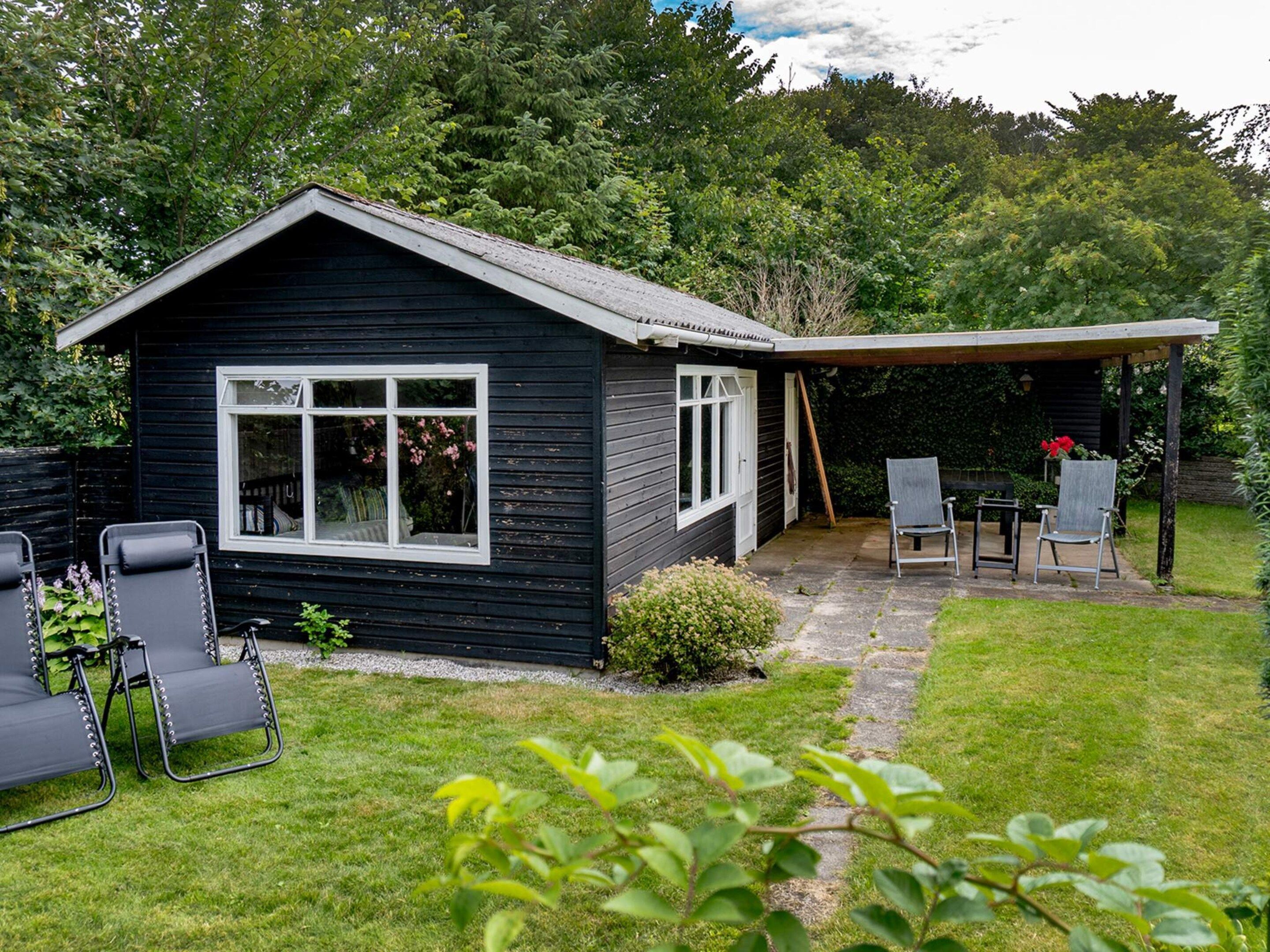 A small black wooden cabin with a covered patio area, two lounge chairs, and a small table.