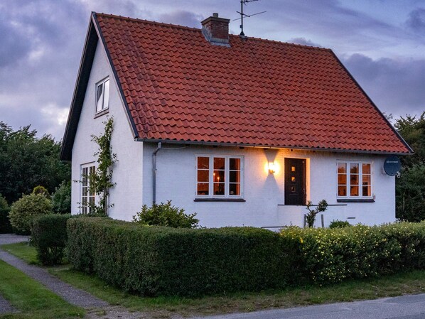 Une maison à deux étages, avec un toit recouvert de tuiles rouges, des murs extérieurs blancs et une porte noire. On peut voir deux fenêtres sur la façade avant, et une antenne parabolique est visible sur le côté droit.