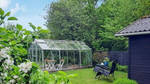 A greenhouse with a table and chairs inside, surrounded by a garden with a wooden shed and a fence. - 6 Person Holiday Home in Bindslev (Bindslev)