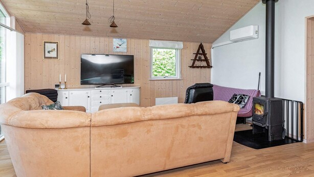 A cozy living room with a large beige sofa, a flat-screen TV, wooden panel walls, a fireplace, and a window with a view of greenery. - 6 Person Holiday Home in Logstor (Logstor)