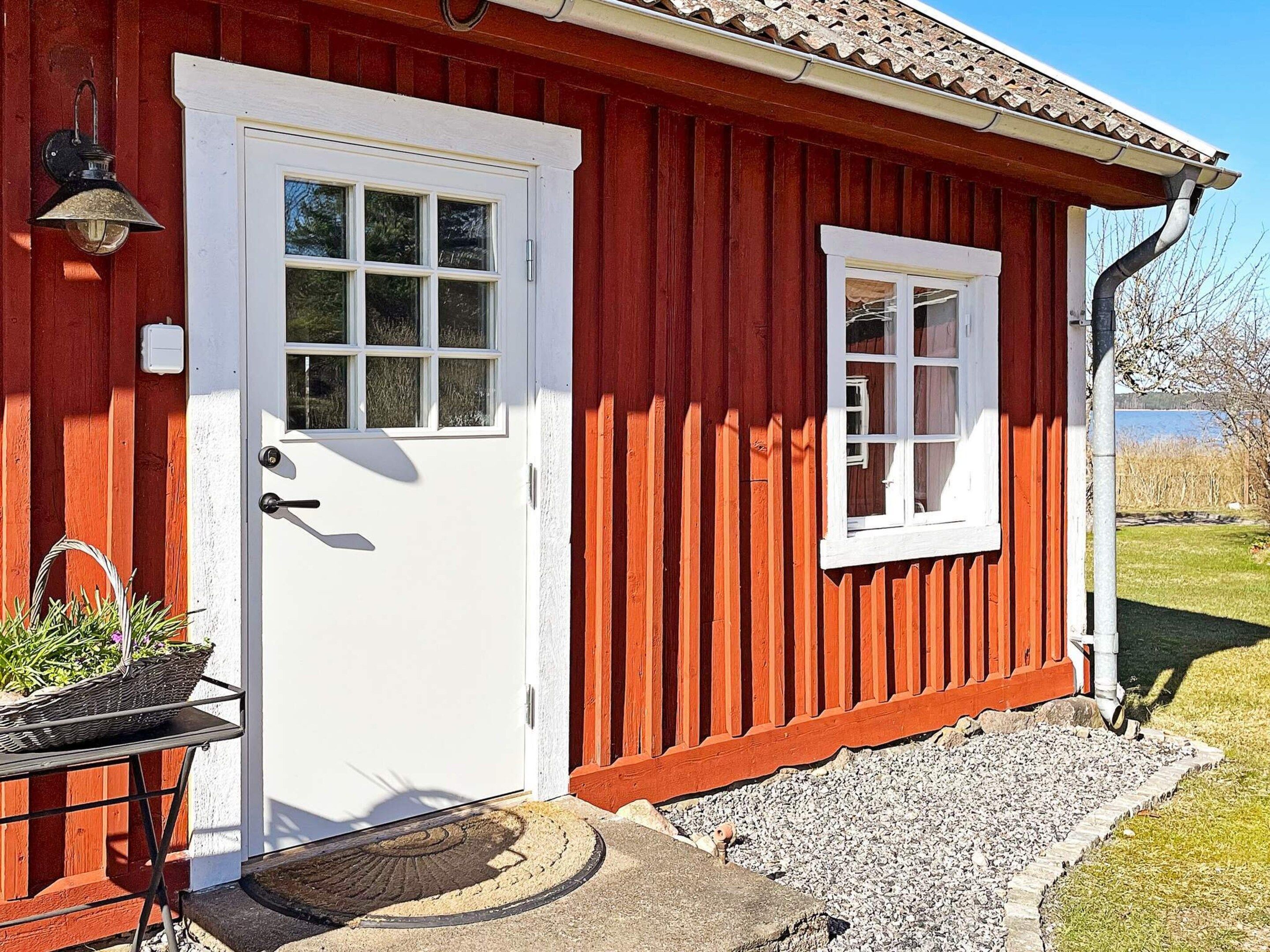 Ein rotes Holzhaus mit weißer Tür und Fenster, einer kleinen Veranda mit Korb und Fußmatte sowie einem Kiesweg, der zur Tür führt.
