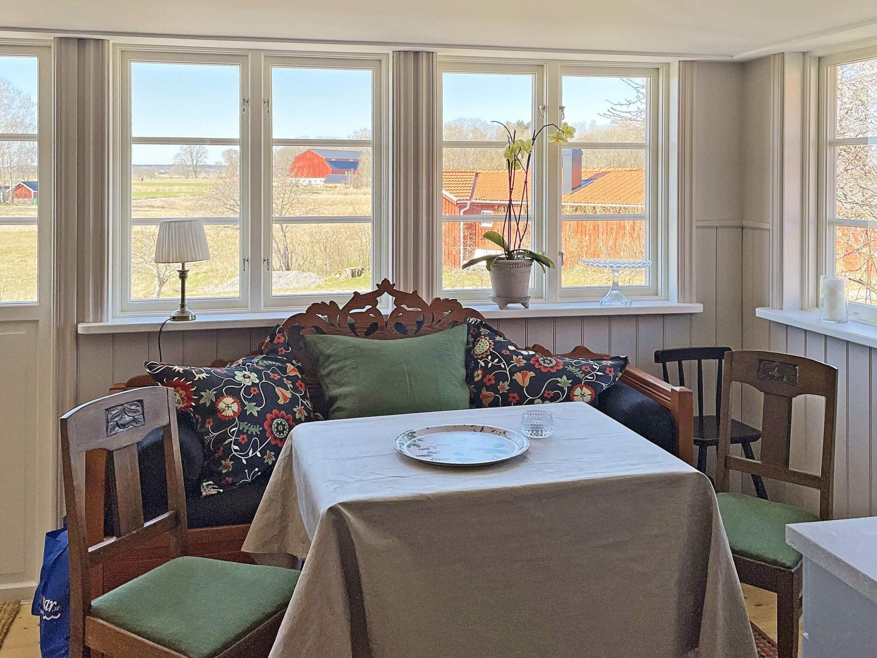 Un comedor con una mesa dispuesta para dos, una ventana con vista a un granero rojo y una planta en maceta en el alféizar.
