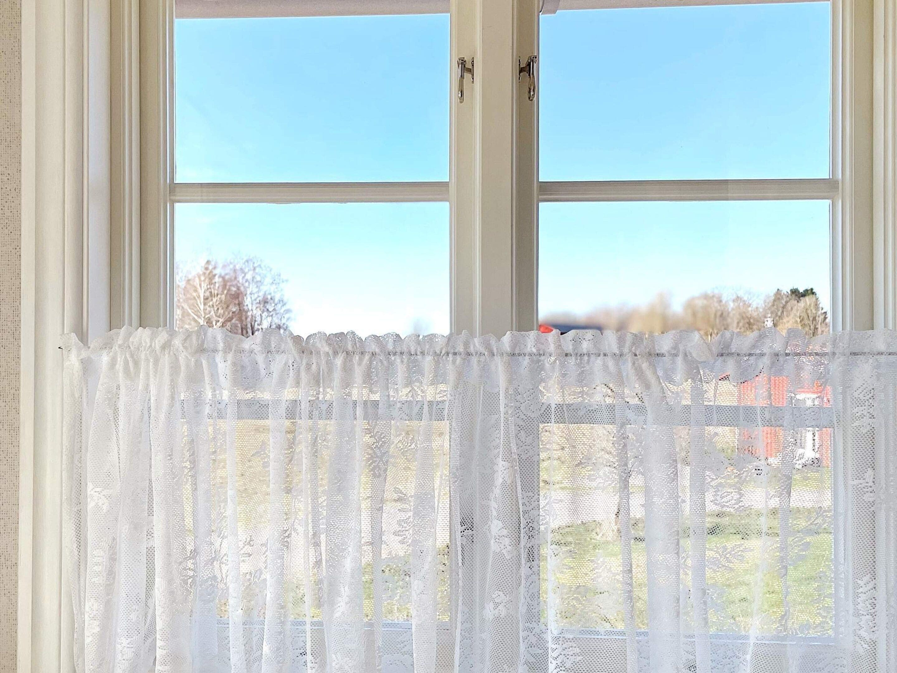 Une fenêtre ornée d’un rideau blanc, légèrement transparent, donnant sur un ciel dégagé et des bâtiments éloignés.
