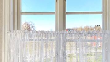 Une fenêtre ornée d’un rideau blanc, légèrement transparent, donnant sur un ciel dégagé et des bâtiments éloignés.