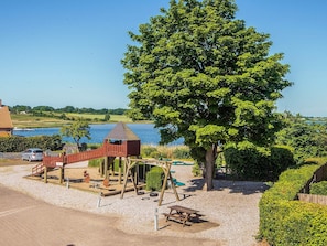 A playground with a swing set and a slide, surrounded by a gravel area and a large tree. - 4 Person Holiday Home in Hejls (Hejls)