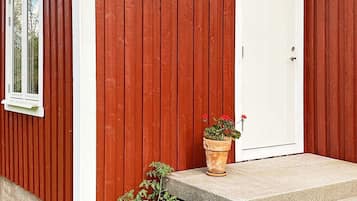 Un edificio de madera roja con puerta blanca y una ventana. Hay un escalón de hormigón que lleva a la puerta, y una maceta con flores rojas en el escalón.