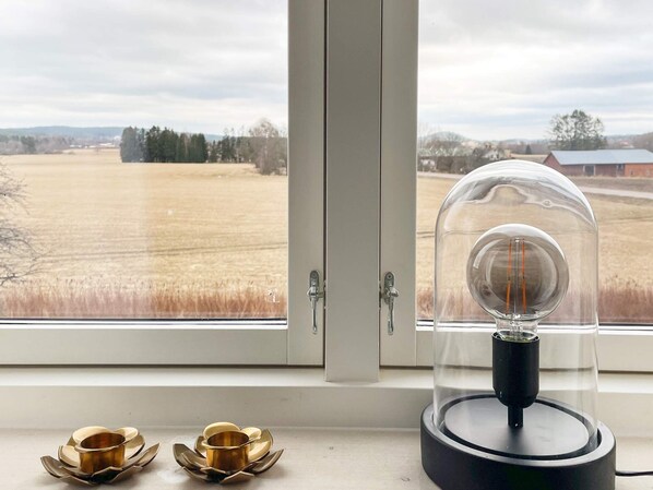 A room with a window overlooking a field, a lamp with a glass dome, and two metallic candle holders on the windowsill. - 4 Star Holiday Home in Vastra Amtervik (Vastra Amtervik)
