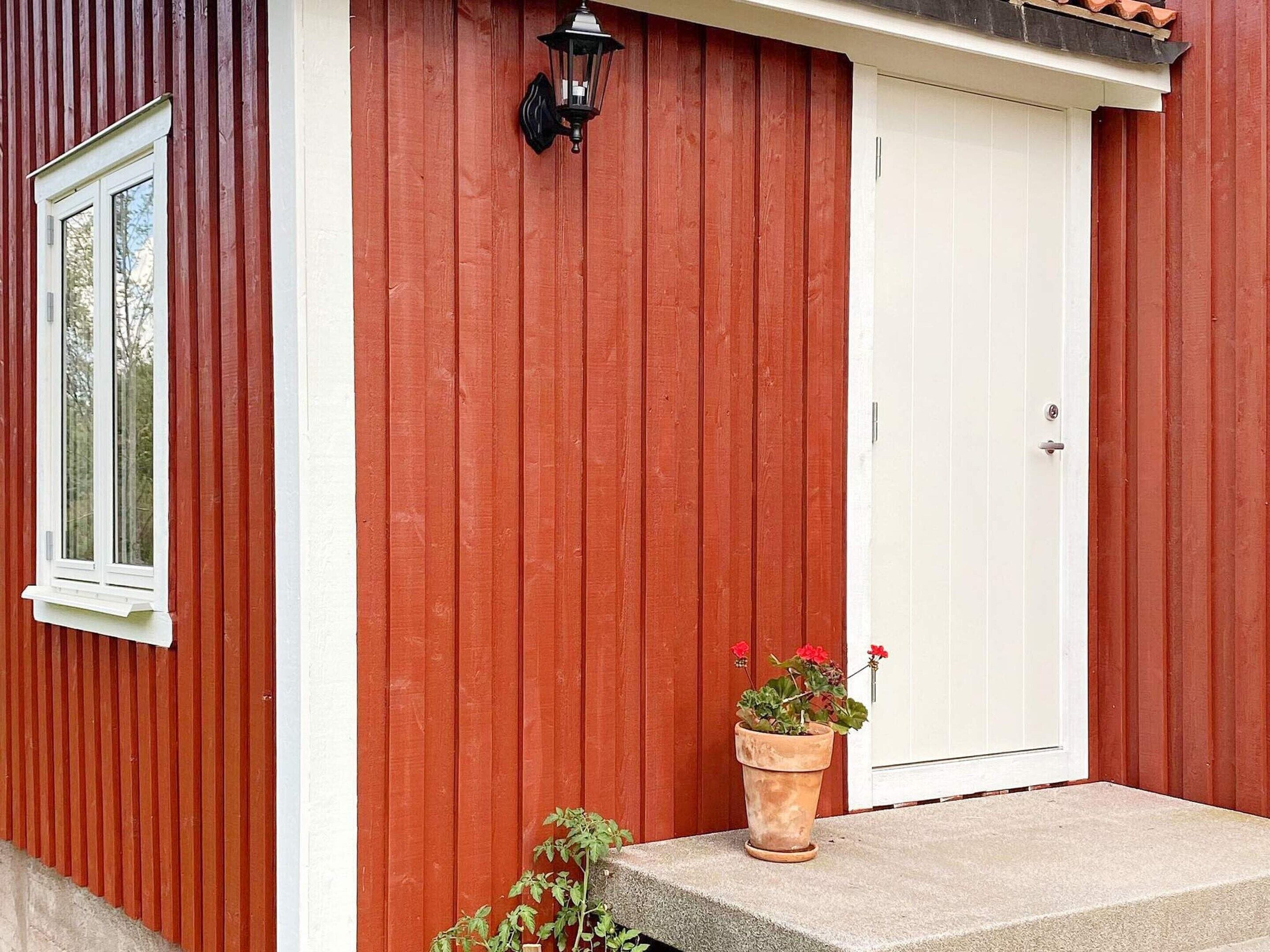 Een rood houten gebouw met een witte deur en een raam. Een potplant met rode bloemen staat op de grond.