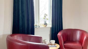 Una habitación con dos sillones de cuero rojo, una mesita redonda con dos tazas y un libro, y una ventana con cortinas.