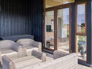 A covered outdoor seating area with wicker furniture, a dark wooden wall, and a sliding glass door leading to a patio. - 8 Person Holiday Home in Store Fuglede (Store Fuglede)