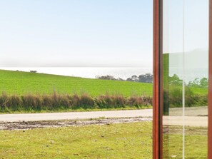 A view of a green field with a dirt path leading to a body of water, viewed through a window with a wooden frame.