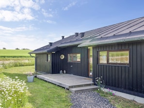 A dark wooden house with a porch, a small garden with flowers, and a clear sky.