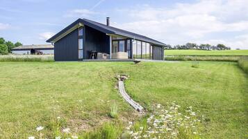 A modern house with a large deck, surrounded by a grassy field and a small stream.